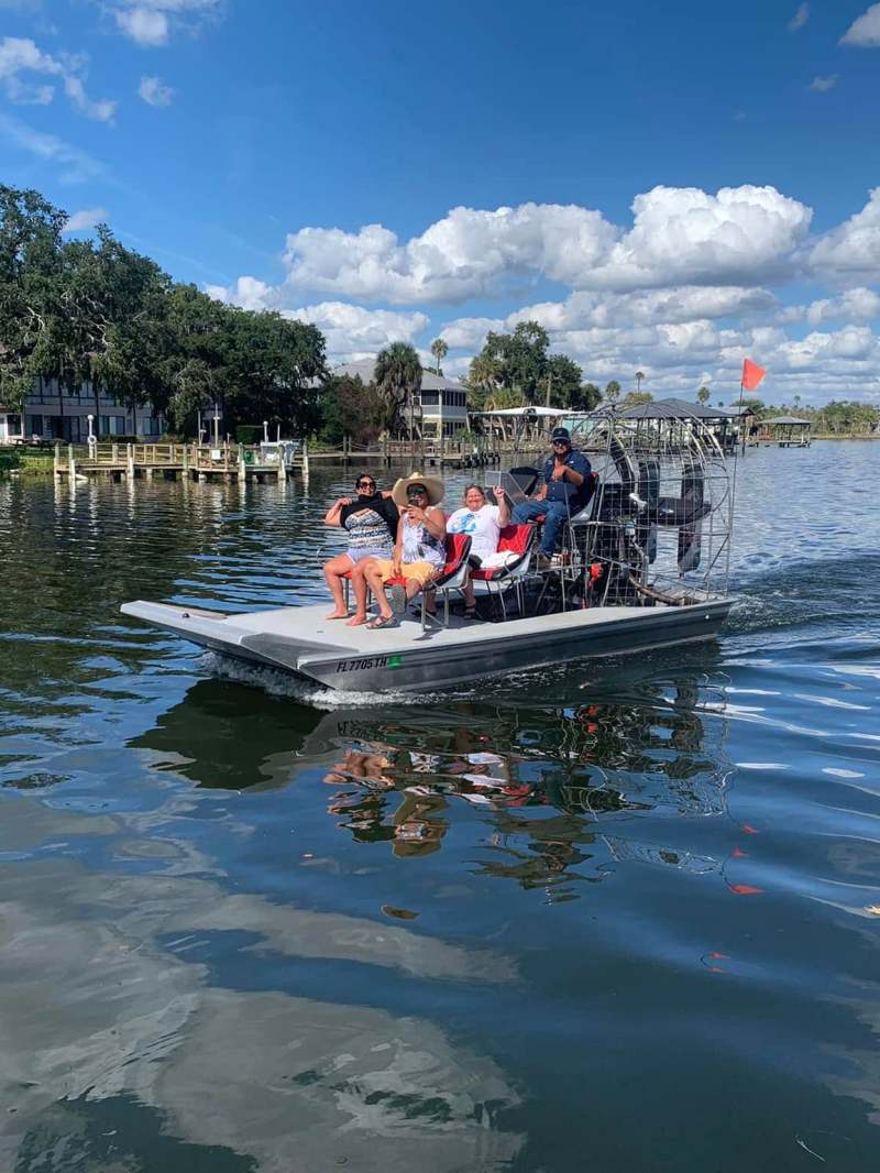 People riding an airboat on a calm lake under a partly cloudy sky.