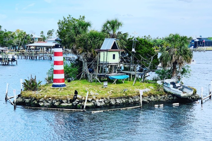 Mini island with a small lighthouse, hut, and boat on a river.