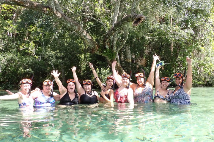 Group of people in swimwear and snorkels standing in clear water, surrounded by trees.