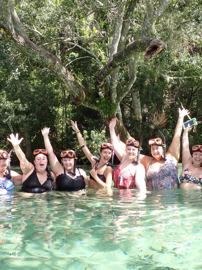 Group of people in swimwear and snorkels standing in clear water, surrounded by trees.