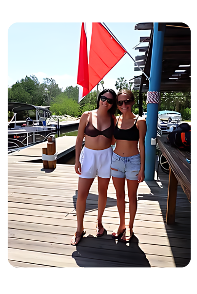 Two women posing on a wooden deck near water, wearing summer attire, with a red flag in the background.