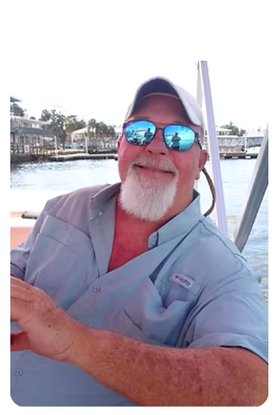 Man with sunglasses and a white beard smiles on a boat, water and buildings in the background.