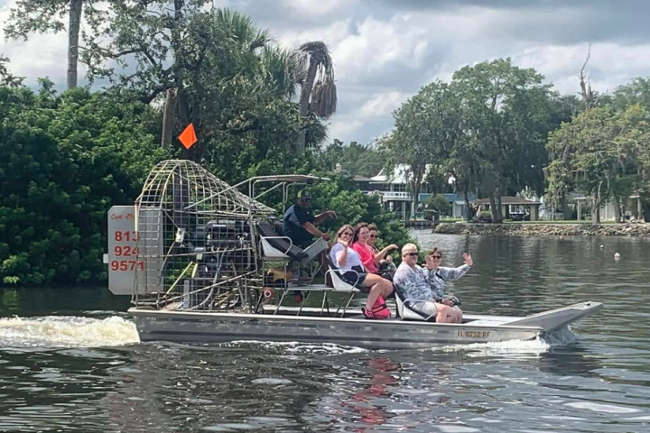 People ride an airboat on a calm river under a cloudy sky.