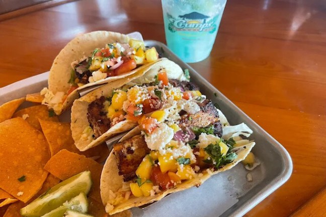 Tray with fish tacos, chips, lime wedges, and a blue drink on a wooden table.