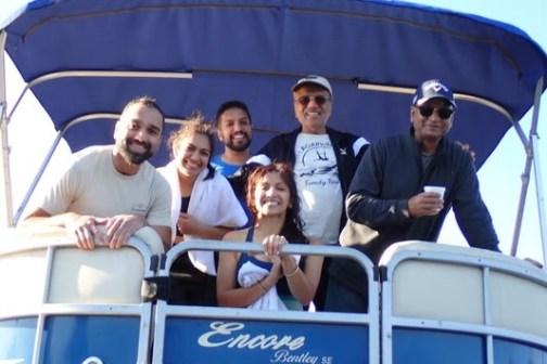 Group of six smiling people on a boat with sunlight and blue canopy.
