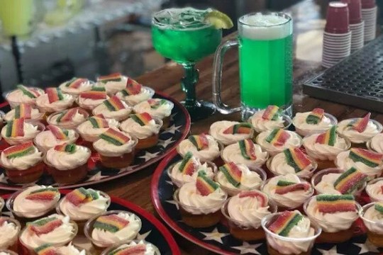 Cupcakes with rainbow candy, green drinks, and cups on a counter.